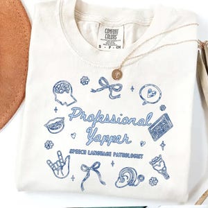 Puede incluir: Camiseta color crema con la frase "Professional Yapper" en azul claro, rodeada de ilustraciones azules. El texto "Speech Language Pathologist" está debajo. Cuello redondo.