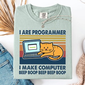 以下が含まれることがあります： 「I ARE PROGRAMMER」の文字と、ノートパソコンとオレンジ色の猫のグラフィックがプリントされた、薄い緑色のTシャツ。「I MAKE COMPUTER BEEP BOOP BEEP BOOP」の文字がグラフィックの下にあります。