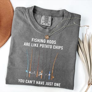 Puede incluir: Una camiseta gris Comfort Colors con el texto "FISHING RODS ARE LIKE POTATO CHIPS YOU CAN'T HAVE JUST ONE". La camiseta presenta un gráfico de varias cañas de pescar en varios colores. Un collar plateado con un colgante está en la camiseta.