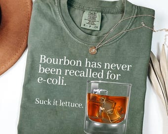 May include: An olive green crew neck t-shirt featuring the white text Bourbon has never been recalled for e-coli. Suck it lettuce. and a graphic of a glass filled with amber liquid and ice cubes. The shirt has a Comfort Colors brand label.