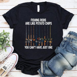 Puede incluir: Camiseta negra con el texto "FISHING RODS ARE LIKE POTATO CHIPS YOU CAN'T HAVE JUST ONE" y un gráfico de cañas de pescar. La camiseta se combina con vaqueros azules y zapatillas blancas.