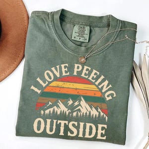 May include: Green Comfort Colors t-shirt with the phrase "I LOVE PEEING OUTSIDE" in white block letters. The design includes a colourful sunset and mountain graphic. A gold necklace and a brown hat are also visible.