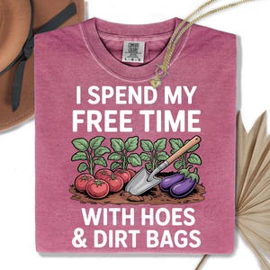 a pink t-shirt with a humorous text and an illustration of gardening tools and vegetables, including tomatoes, eggplants, and a shovel.