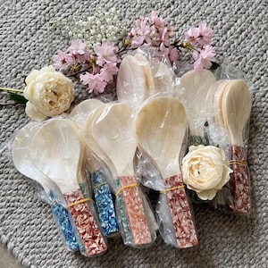 May include: A selection of rice spoons with pearlescent bowls and patterned handles in red, blue, and peach. The spoons are packaged in clear plastic. The handles are decorated with a mosaic pattern. Flowers are arranged around the spoons.