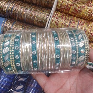 Puede incluir: Un conjunto de pulseras plateadas con detalles turquesa y plateados, presentado en un embalaje de plástico transparente. Las pulseras tienen bandas plateadas lisas, bandas de purpurina turquesa y bandas adornadas con pedrería transparente y adornos plateados. Se ven otras pulseras en el fondo.