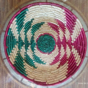May include: A round, woven basket with a spiral design. The basket features alternating sections of green, white, and pink, with a beige center. The rim is a darker pink. The basket is likely used for decorative or storage purposes.