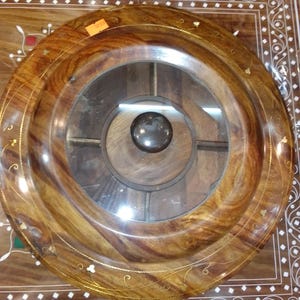 May include: A round, wooden decorative tray with a clear glass center and a dark, spherical knob. The tray has a rich, brown wood grain with gold-colored accents and is set on a decorative wooden surface.