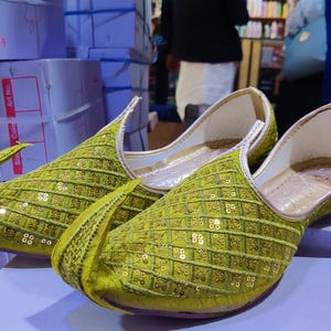 Puede incluir: Un par de zapatos verdes sin cordones con lentejuelas. Los zapatos tienen una punta afilada y un patrón decorativo. El interior es blanco y los zapatos se exhiben sobre una superficie de color púrpura claro. El fondo incluye cajas y personas.