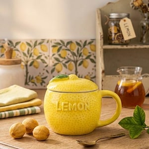 Könnte beinhalten: Eine gelbe Keramiktasse in Zitronenform mit passendem Deckel und dem geprägten Wort "LEMON" auf der Vorderseite. Die Tasse hat einen Henkel und eine strukturierte Oberfläche. Der Hintergrund umfasst eine Holzoberfläche, einen Löffel und einen kleinen Krug.
