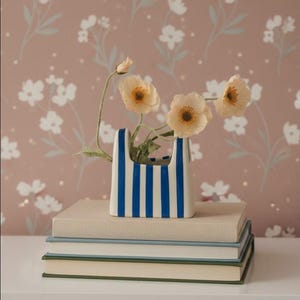 Puede incluir: Un jarrón de cerámica blanco con rayas verticales azules contiene varias flores de color melocotón. El jarrón está sobre una pila de libros con un fondo de pantalla floral. Las flores tienen centros amarillos.