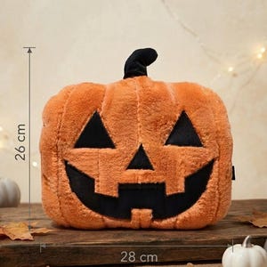 Op de afbeelding: Een oranje pluche pompoen met een zwarte steel en een zwart jack-o'-lantern gezicht met driehoekige ogen en een brede, grillige mond. De pompoen is 26 cm hoog en 28 cm breed, zoals aangegeven door de zichtbare meetmarkeringen op de afbeelding.