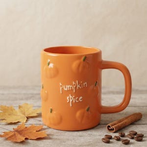 Op de afbeelding: Een oranje keramische mok met een herhalend reliëfpatroon van pompoenen en de witte tekst "pumpkin spice" op de voorzijde. De mok heeft een gebogen handvat en een gladde afwerking, geschikt voor het drinken van warme dranken in de herfst.