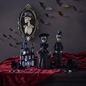 Può includere: Due statuette di scheletri vestite con abiti formali neri, una con un cappello a tesa larga e l'altra con un cilindro, accanto a una casa stregata in miniatura nera con accenti di fantasmi bianchi e un ritratto di scheletro incorniciato.