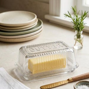 Könnte beinhalten: Eine Butterdose aus klarem Glas mit einem rechteckigen Deckel, auf dem das Wort BUTTER eingeprägt ist. Das Gefäß hat eine gerippte Struktur und enthält ein Stück gelbe Butter. Der Deckel sitzt auf einem passenden Glasunterteil.