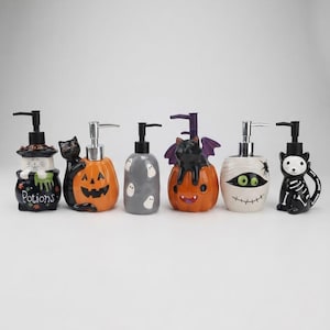 Pode incluir: Um conjunto de seis dispensadores de sabonete de cerâmica com temas de Halloween, incluindo um gato preto com um caldeirão com a inscrição Potions, um gato com abóbora, uma garrafa de fantasma cinzenta, um gato com asas de morcego numa abóbora, uma múmia e um gato esqueleto.