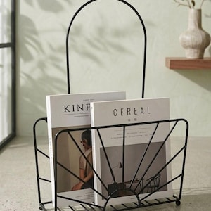 Op de afbeelding: Een zwarte metalen tijdschriftenhouder met een draadframe in de vorm van een zonnestraal en een hoge, gebogen handgreep. De houder bevat twee tijdschriften, getiteld Kinfolk en Cereal, die rechtop in de draadstructuur zijn geplaatst.