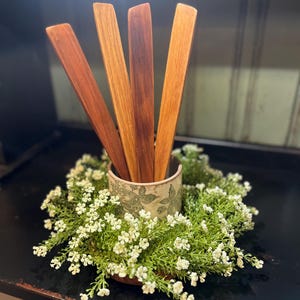 May include: Four wooden spoons in a white ceramic utensil holder with a floral design. The holder is surrounded by a green wreath with white flowers.