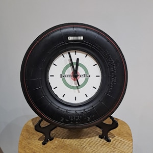 May include: A circular clock made from a black tyre, featuring a red trim. The clock face is white, displaying the word "Lambretta" and a green and red target. The clock is supported by a black metal stand.