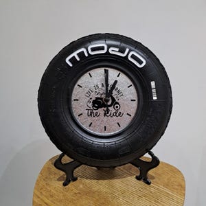 May include: A black tyre clock with the word "MOJO" in white lettering. The clock face is silver with black hands and the text "Life is a Journey, Enjoy The Ride". The clock is displayed on a black metal stand.