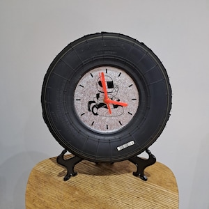 Pode incluir: Um relógio de pneu preto com tema de corrida. O mostrador do relógio apresenta uma ilustração em preto e branco de um piloto de kart com ponteiros vermelhos. O relógio é montado em um suporte preto e fica em uma superfície de madeira.