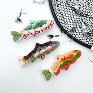 Puede incluir: Tres arreglos de caramelos con forma de pez llenos de caramelos de goma de colores. Los peces son blancos con detalles de caramelos verdes, rojos, marrones y naranjas. También hay señuelos de pesca y una red de pesca negra en la imagen.