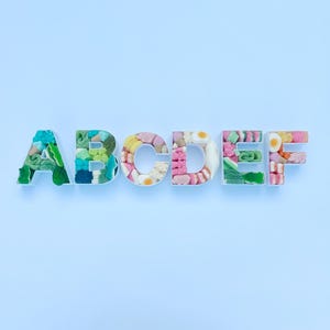 Könnte beinhalten: Die Buchstaben A, B, C, D, E und F sind mit bunten Süßigkeiten gefüllt. Die weißen Buchstaben heben sich von einem hellblauen Hintergrund ab. Die Süßigkeiten umfassen grüne, rosa, gelbe und weiße Süßigkeiten.