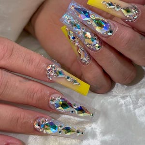 Könnte beinhalten: Lange, gelbe und transparente Acrylnägel mit schillernden Strasssteinen und Edelsteinen. Die Nägel sind mit Kristallen in verschiedenen Formen und Größen verziert, wodurch ein glamouröses Design entsteht. Die Nägel haben eine Stiletto-Form.