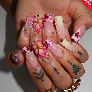 Könnte beinhalten: Eine Hand mit langen, verzierten Kunstnägeln. Die Nägel weisen verschiedene Designs auf, darunter rosa und weiße French-Spitzen, florale Verzierungen und Perlenakzente. Einige Nägel haben einen 3D-Effekt. Ein rot-weißer "50% OFF"-Aufkleber befindet sich in der oberen rechten Ecke.