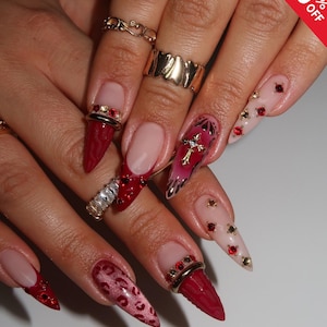Peut inclure: Gros plan sur des ongles manucurés avec divers motifs. Certains ongles sont rouge foncé avec des accents dorés et rouges, d'autres sont nude avec des embellissements rouges et dorés. Un ongle présente une croix.