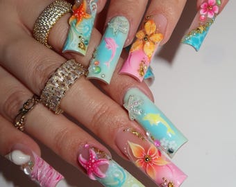 May include: Long square-shaped artificial nails featuring a tropical theme with gradient blue and pink backgrounds. The nail art includes three-dimensional starfish, dolphins, and floral designs, accented with small gold beads and clear rhinestones.