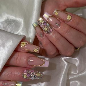 May include: Long, coffin-shaped acrylic nails with a light pink base and gold accents. Some nails feature small yellow flowers, while others are adorned with clusters of colorful rhinestones and gems. The nails have a glossy finish.