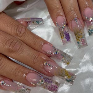 May include: Long, square-shaped acrylic nails with a clear overlay, featuring dried flowers and rhinestones. The nails have a pink base with a French tip design. The design includes yellow, purple, and white flowers.