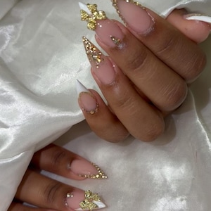 May include: Close-up of manicured nails with a French tip design. The nails are long and pointed, with a nude base and white tips. Gold embellishments, including rhinestones and bow-shaped charms, adorn the nails, creating a glamorous look.