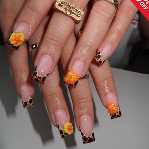 May include: Close-up of a hand with long, square acrylic nails. The nails have a French tip design with a leopard print pattern. Each nail features a 3D orange and yellow flower. Gold rings are also visible on the fingers.