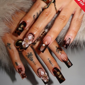 May include: A close-up of a hand with long, square-shaped acrylic nails. The nails feature various designs, including floral accents, tortoiseshell patterns, and dark brown tips. Gold rings and a decorative gold object are also visible.
