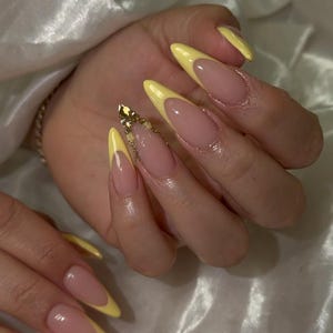 Puede incluir: Uñas largas en forma de almendra con diseño de punta francesa amarilla. Las uñas tienen una base nude y están adornadas con adornos dorados en una uña. Las uñas están bien cuidadas y pulidas.