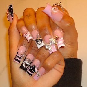 Op de afbeelding: Lange vierkante kunstnagels met een French manicure basis met roze en zwarte accenten. De ontwerpen bevatten stippen, horizontale strepen, een hartvormige hangslotbedel, een Hello Kitty-figuurtje en diverse strass-steentjes als versiering.