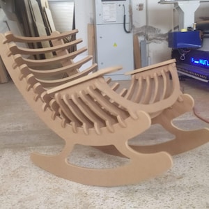 May include: A light brown wooden rocking chair with a unique, open-frame design. The chair features a curved back and rockers, constructed from layered wooden slats. The chair is in a workshop setting.