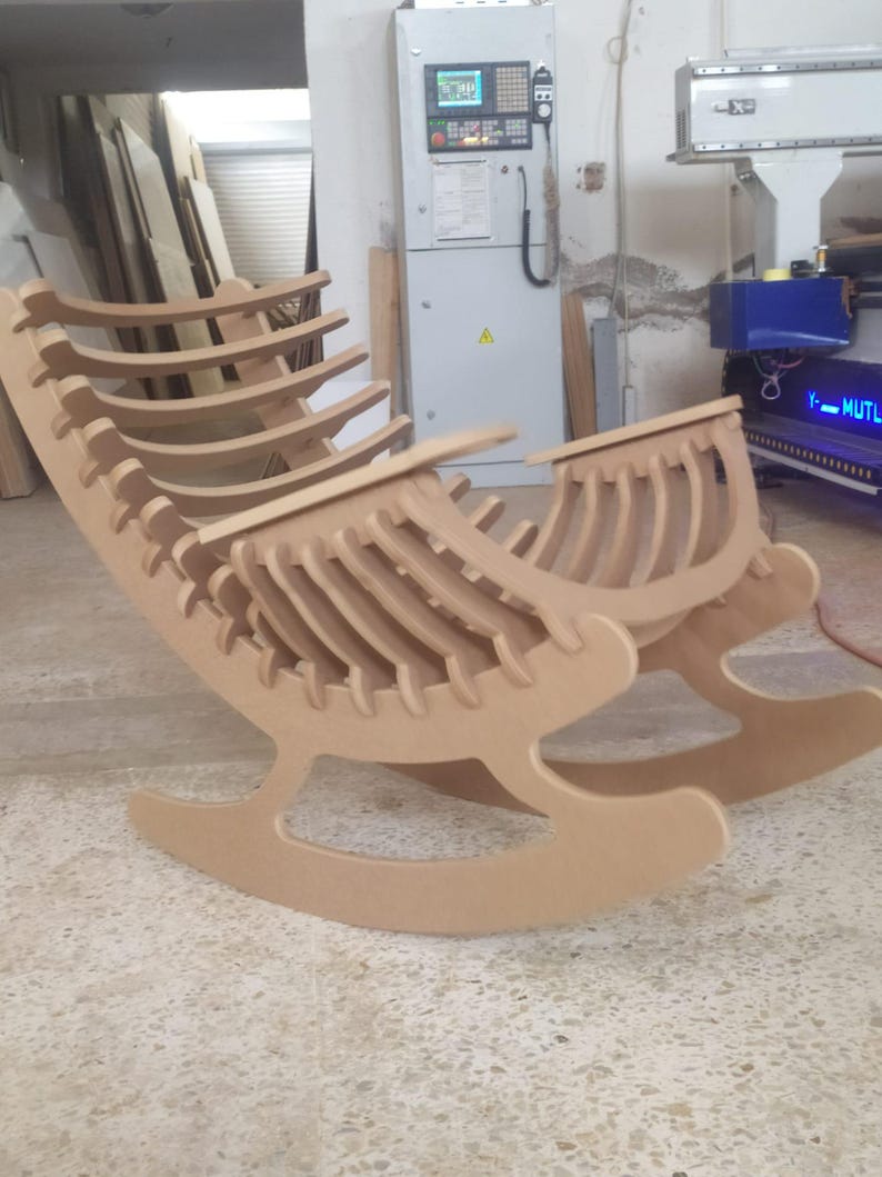 May include: A light brown wooden rocking chair with a unique, open-frame design. The chair features a curved back and rockers, constructed from layered wooden slats. The chair is in a workshop setting.
