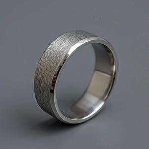 Op de afbeelding: Een zilverkleurige ring met een getextureerde band en gepolijste randen. De ring heeft een eenvoudig, klassiek ontwerp en staat tegen een grijze achtergrond. De ring is ongeveer 6 mm breed, of 0,6 cm.