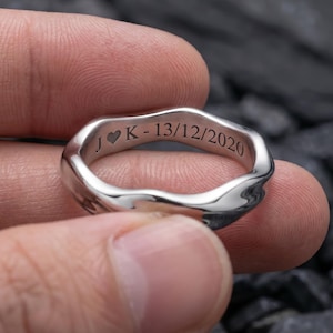 May include: A silver-colored ring with a wavy design. The ring has engraved text: a heart symbol, the letter "K", and the date "13/12/2020". The ring is held between fingers against a dark background.
