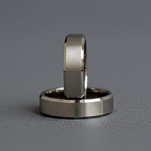 May include: Two metallic rings with a brushed finish and beveled edges. One ring is positioned horizontally at the base, while the other ring stands vertically upright behind it, showcasing a flat band profile and a neutral gray color tone.