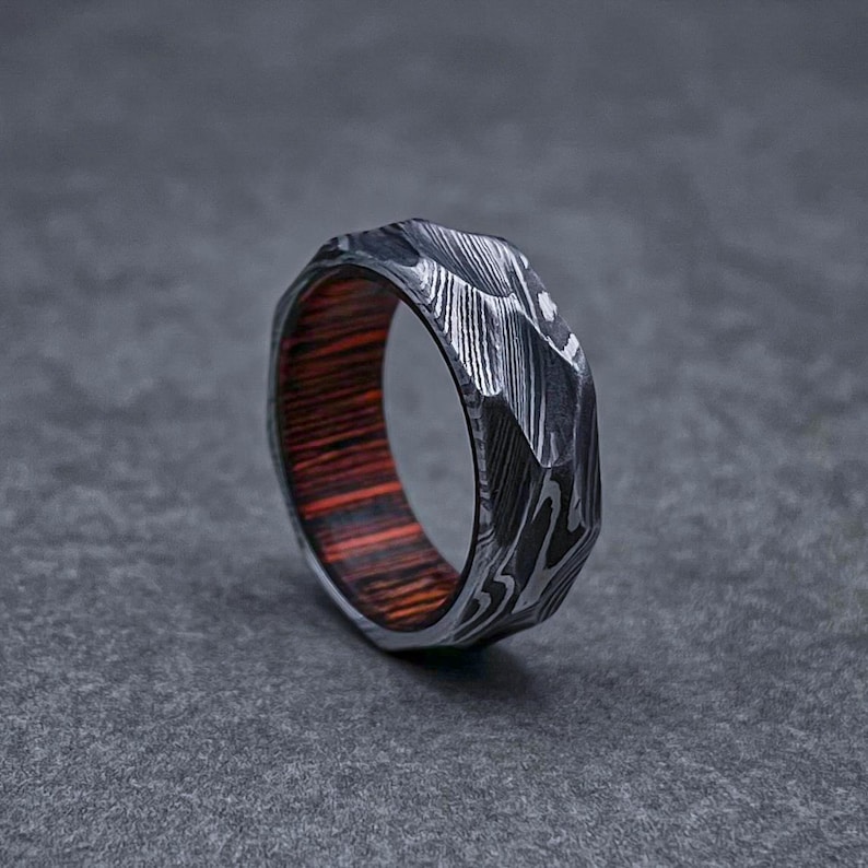 May include: A silver ring with a textured, faceted design. The ring has a wooden inlay with red and brown stripes.