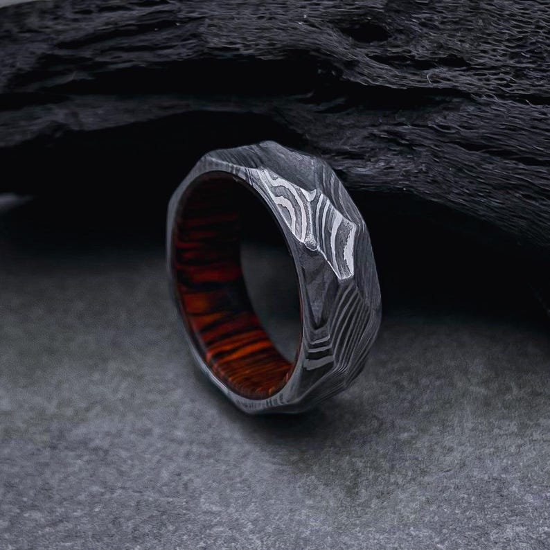 May include: A silver ring with a textured, patterned design and a red wood inlay. The ring is on a dark gray surface.