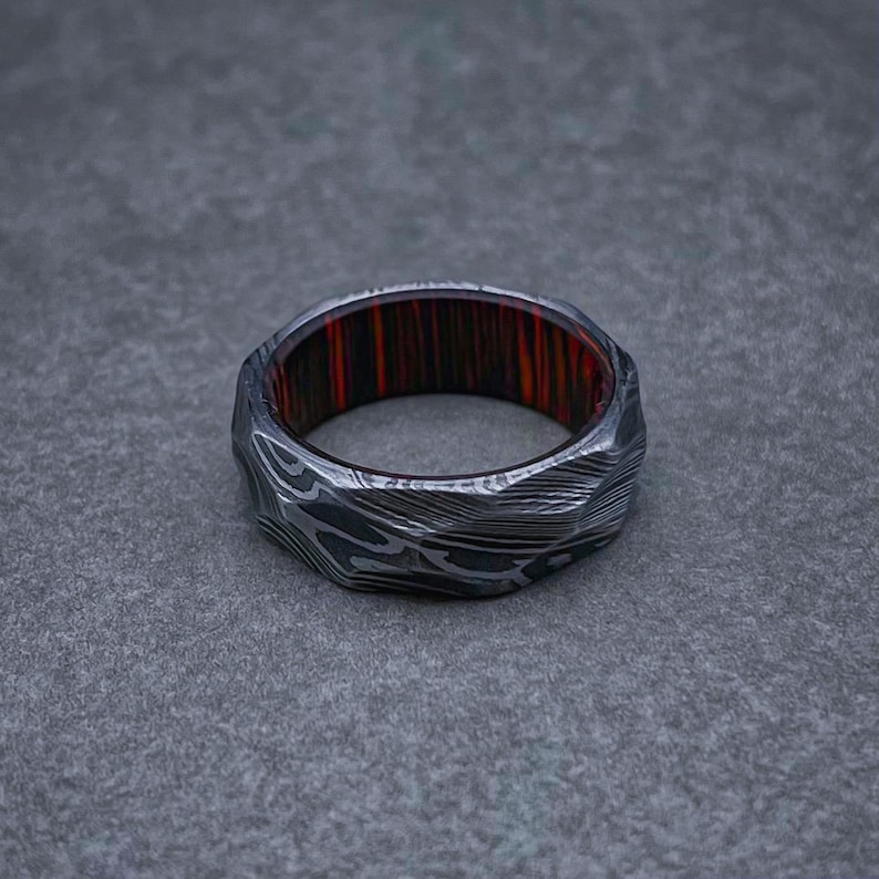 May include: A silver ring with a textured, patterned surface and a red and black wood inlay.
