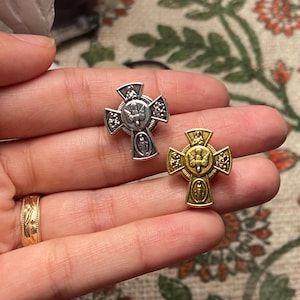 Puede incluir: Se muestran dos prendedores religiosos con forma de cruz, uno plateado y otro dorado. Cada cruz presenta una paloma, una figura y detalles ornamentados. Los prendedores se sostienen en una mano, mostrando su tamaño y diseño. El prendedor dorado tiene un tono ligeramente más oscuro.