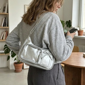 Peut inclure: Sac à bandoulière argenté avec une finition brillante. Le sac comporte deux poches avant et un nœud décoratif. La bandoulière est également argentée. Un accessoire de mode.