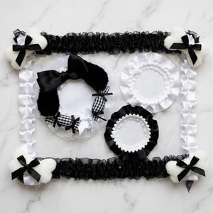 Peut inclure: Un cadre rectangulaire en tissu blanc à volants avec des nœuds noirs sur des coins en peluche blanche en forme d'os. À l'intérieur se trouvent trois accessoires circulaires : un avec de la fourrure noire et des accents vichy, un cercle blanc à volants et un cercle noir à volants.