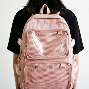 Peut inclure: Un sac à dos rose avec plusieurs compartiments et des poches en filet. Le sac à dos a une poignée supérieure, des bretelles réglables et des boucles blanches. L'avant comporte deux poches en filet transparentes.