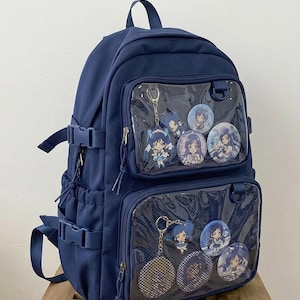 Könnte beinhalten: Marineblauer Rucksack mit transparenten Fronttaschen, die Schlüsselanhänger und Buttons mit Anime-Charakteren zeigen. Der Rucksack hat verstellbare Träger, Schnallen und einen oberen Griff. Hergestellt aus strapazierfähigem Material.
